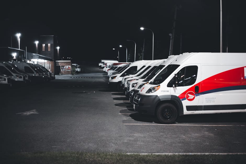 Nighttime view of a parking lot with a row of white delivery vans featuring red and blue branding, parked side by side on a paved surface illuminated by tall streetlights. The vans are positioned near