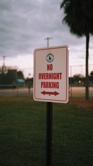 A rectangular traffic sign with a white background and red border is mounted on a metal post, indicating no parking at any time with a bold black 'P' crossed out by a red circle and slash. Below the s