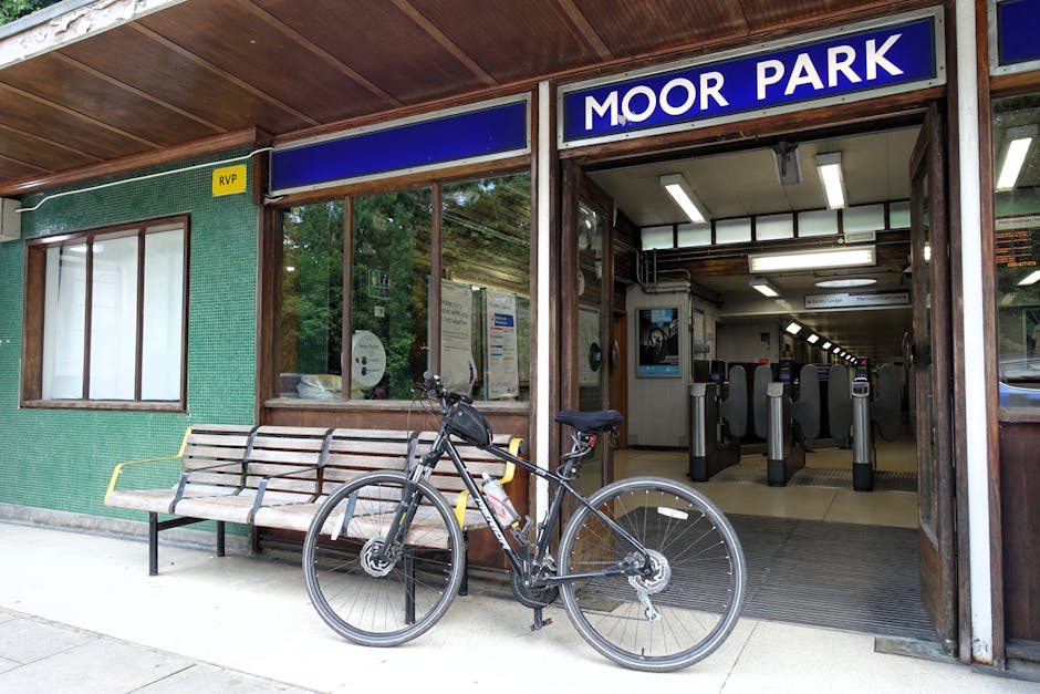 A photograph of the exterior of Moor Park station showing the entrance with a blue sign displaying 'MOOR PARK' above the ticket gates. To the left of the entrance, there is a wooden bench with metal armrests and a black bicycle parked in front of it, secured to the bench with a lock. The station building features large glass windows revealing the interior, where ticket barriers and a ticket machine are visible. Inside, fluorescent lighting illuminates the area, and a few posters or notices are displayed on the walls. The building's exterior wall is covered with green tiles, and a small yellow sign marked 'RVP' is attached near the window. The scene is set on a paved pavement, and the overall environment appears clean and well-maintained, typical of a location where house removals and furniture transport logistics may take place in connection with a home relocation service provided by Man With a Van Crofton Park.