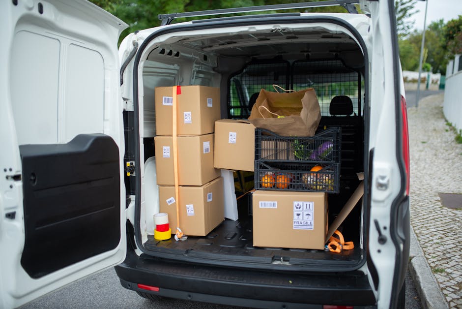 The rear cargo area of a white van is filled with various packed moving boxes wrapped in brown packing paper, some with barcode labels, stacked and arranged to optimise space. A black plastic crate contains fresh produce such as oranges and vegetables, positioned among the boxes. A brown paper shopping bag also sits among the boxes, possibly containing additional items to be packed or carried during the home relocation process. The van is parked on a paved street near a white fence, with a glimpse of green foliage and trees in the background, indicating an outdoor setting. Inside the van, a black cargo barrier separates the loading area from the driver’s cabin, and a rolled-up carpet or foam pipe is visible at the side. There are packing materials like cardboard and plastic wraps present, along with a small yellow and red item resembling a protective cap or label. The scene captures the loading process involved in furniture transport or packing and moving services conducted by Man With a Van Crofton Park, showcasing a typical house removal preparation.