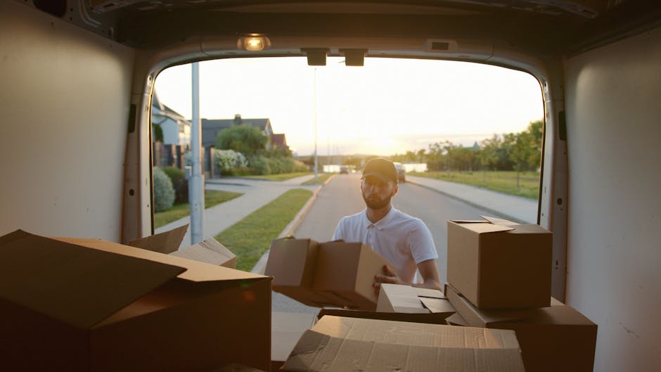 A man inside a moving van, seen through its open rear doors, loads a cardboard box while surrounded by several other cardboard boxes and flat-pack furniture pieces, some wrapped in plastic for protect