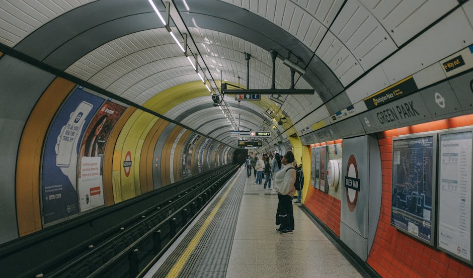 Inside the Green Park London Underground station, several passengers stand on the platform preparing for travel, with some engaging with their phones or waiting near the yellow and orange tiled walls. The curved ceiling of the underground station is lined with bright lighting, and digital information screens display train departure times and routes. The station features safety and directional signage, including the London Underground roundel sign with the station name. The platform has a yellow safety line along the edge, and advertisements are mounted on the walls. This station scene provides a context for a home relocation or moving service, such as those offered by Man With a Van Crofton Park, especially when considering transportation logistics related to house removals or furniture transport from this underground station to a new residence or storage facility.
