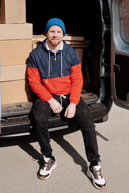 A man wearing a black cap and light-colored T-shirt is inside a van, carefully handling a medium-sized cardboard box during a home relocation. He is stacking additional boxes of varying sizes, all sea
